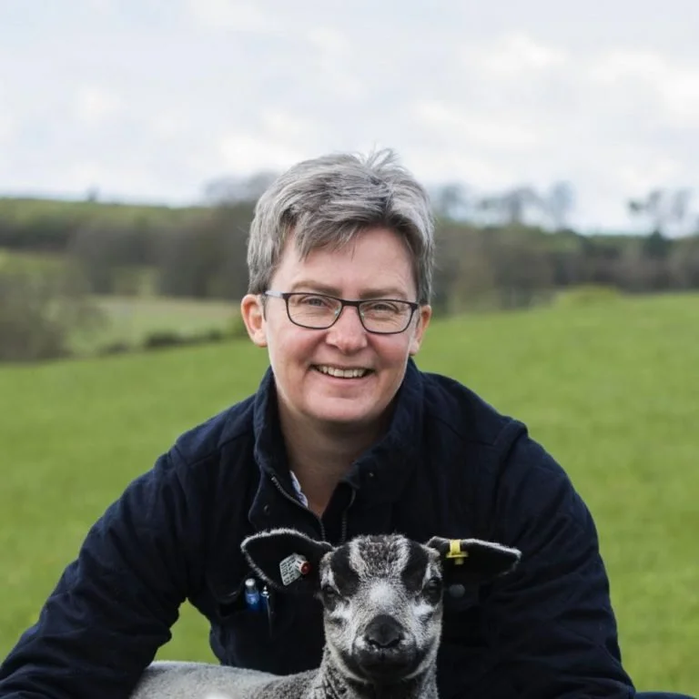 A person with short gray hair, glasses, wearing a navy jacket, smiling outdoors in a green field with a dog, a black and white puppy, in front of them on a cloudy day.