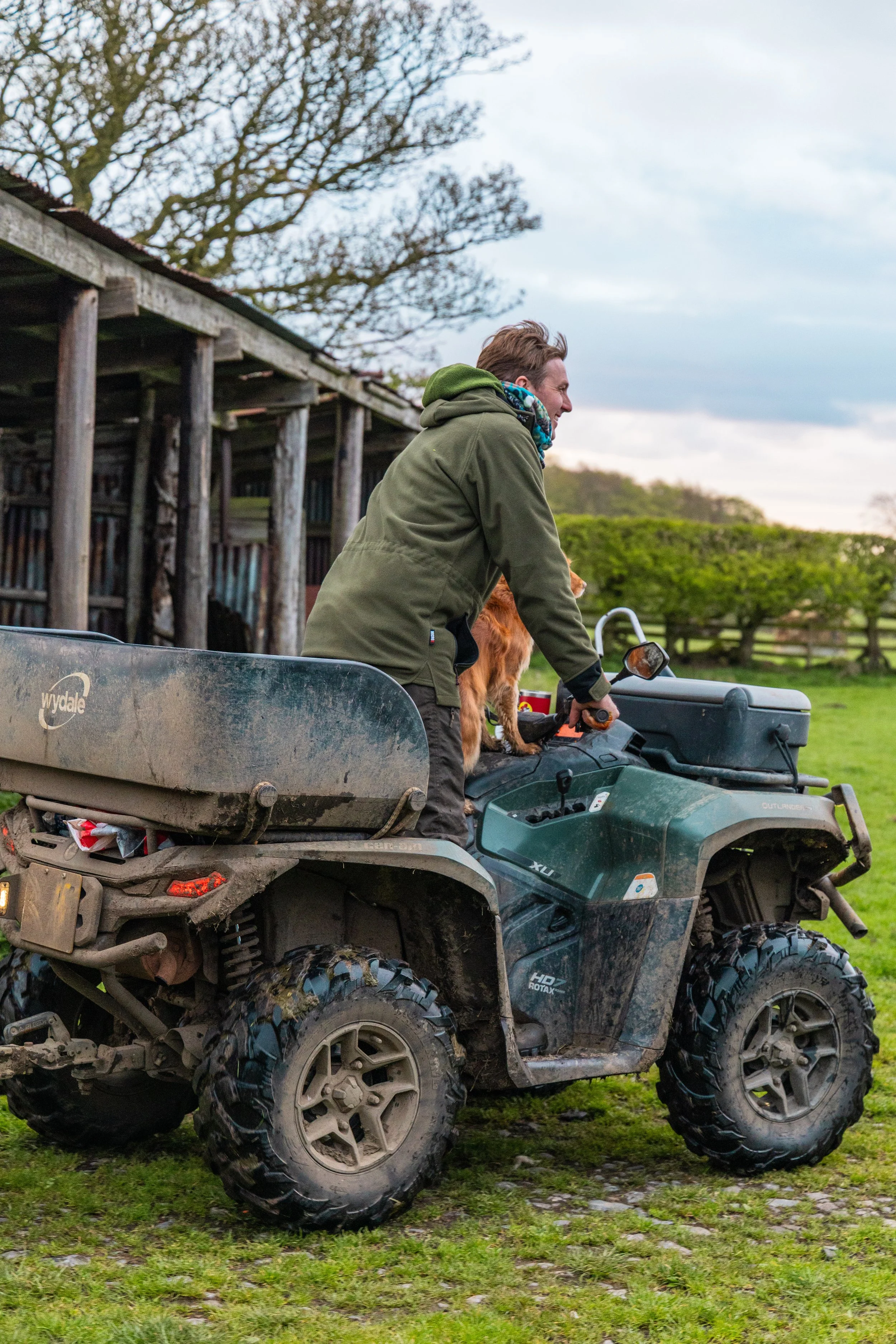 A man riding a ATV in a grassy field with a dog sitting on his lap. The man is smiling and wearing a green jacket and a multicolored scarf. There are trees and a wooden structure in the background.