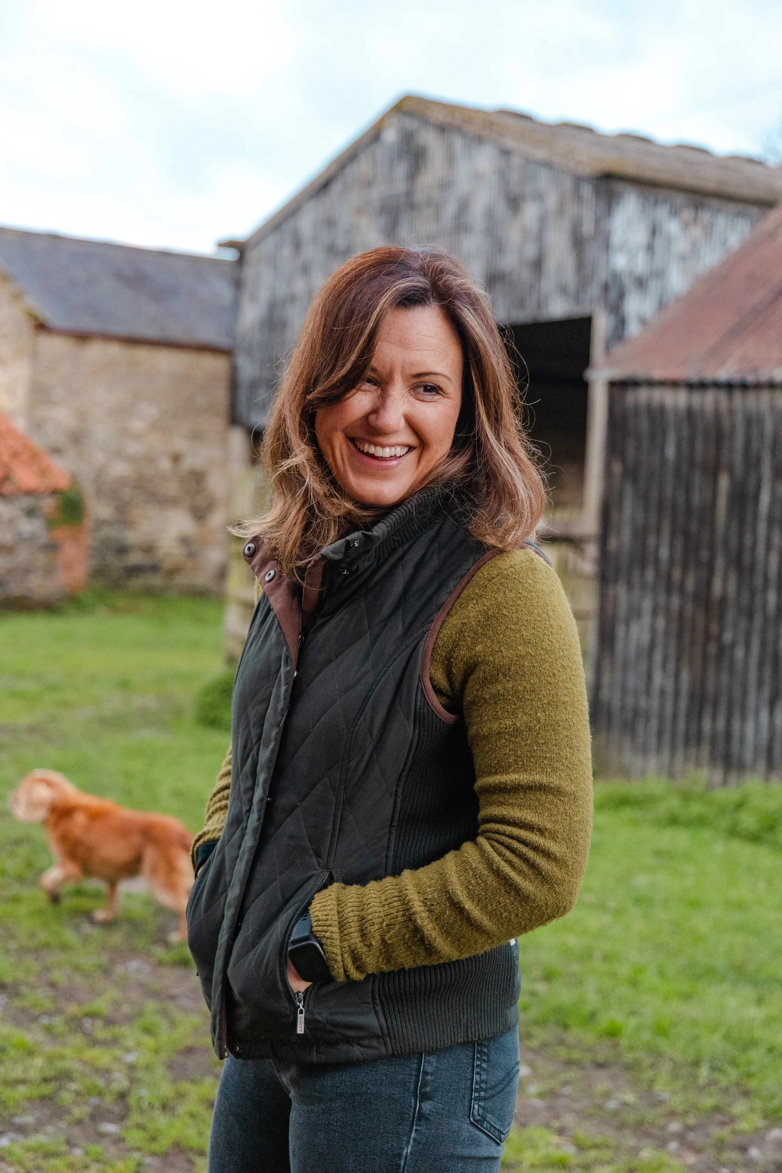 A woman smiling outdoors with a barn and a dog in the background