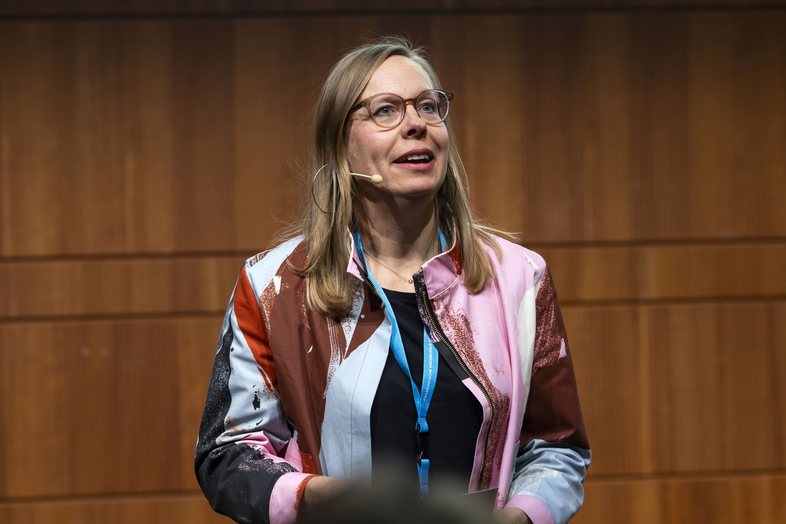 Frau mit langen Haaren, Brille und einem Microphone auf einer Bühne vor einer Holzwand.