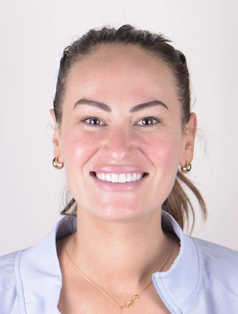 A woman with light skin and brown hair pulled back, smiling at the camera, wearing a blue collared shirt and gold jewelry.