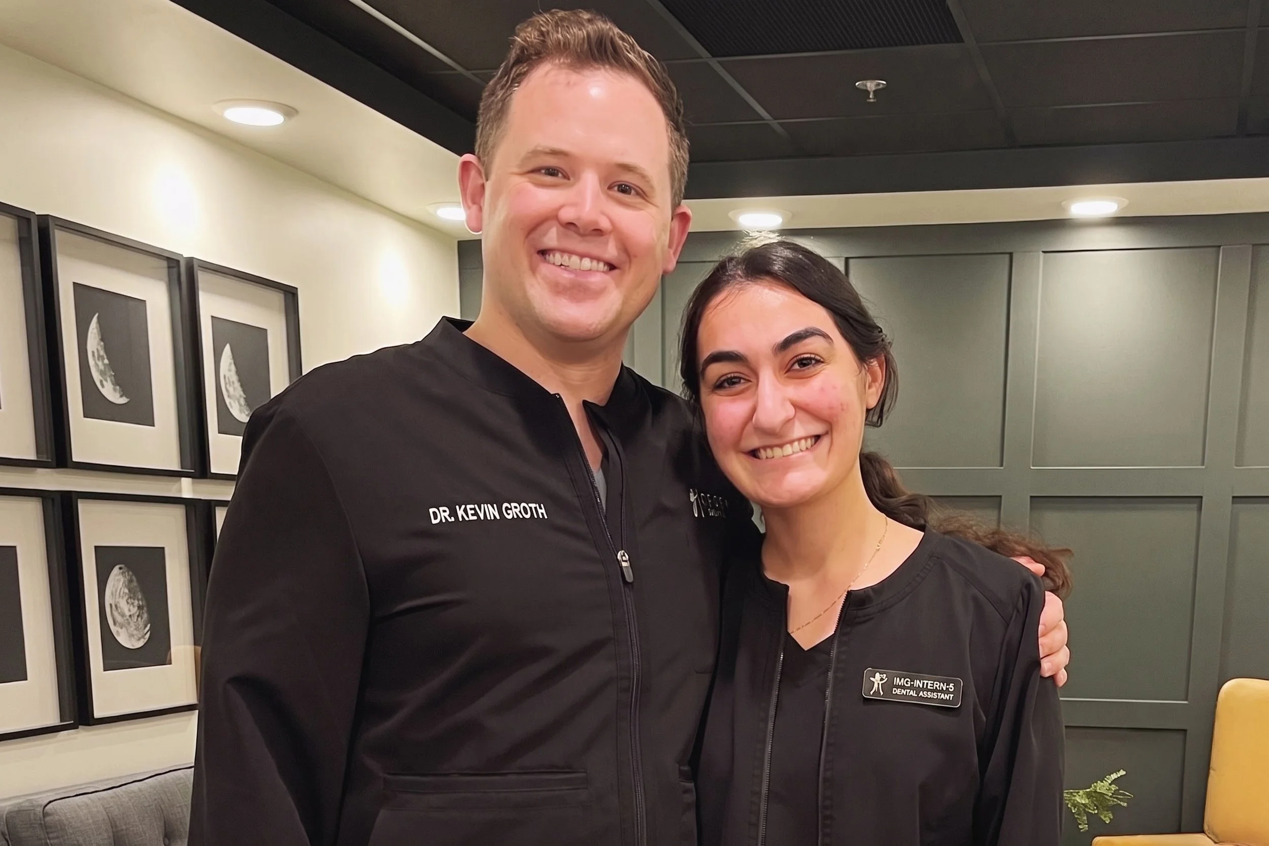 A smiling man and woman in black dental scrubs standing close together indoors. The man has short, light brown hair and is wearing a jacket with the name tag of Dr. Kevin Groth. The woman has dark brown hair tied back and a badge indicating she is a 