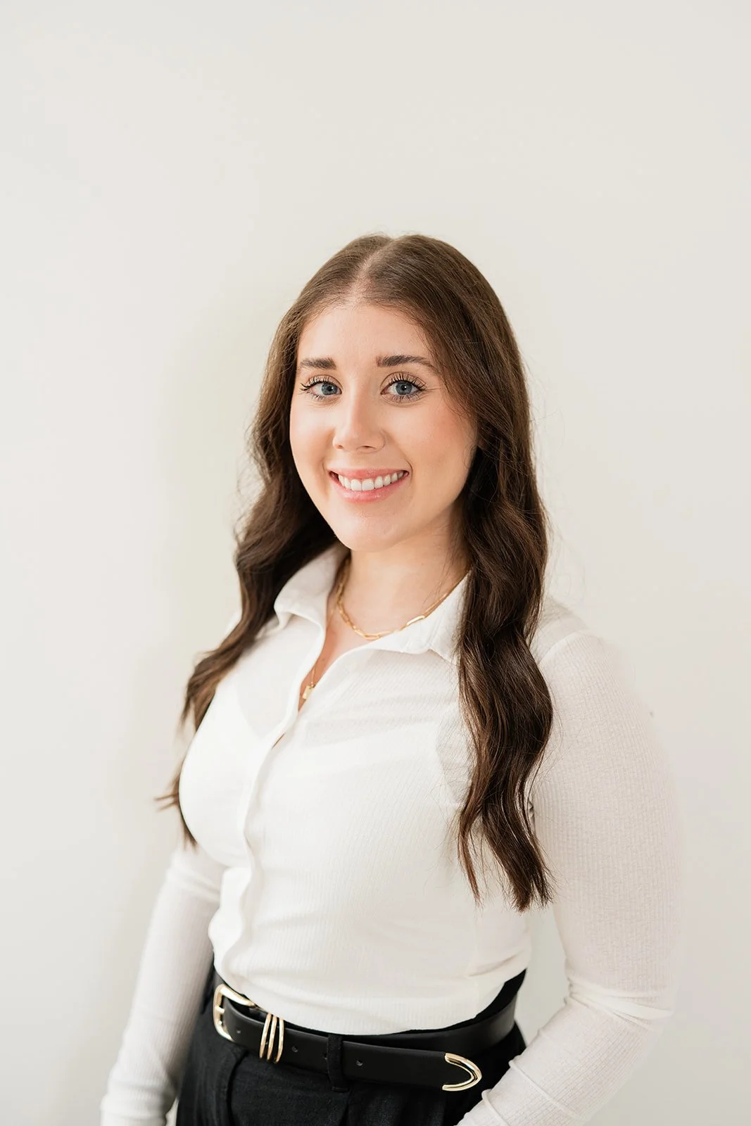 Portrait of a young woman with long brown hair, blue eyes, wearing a white blouse and black belt, smiling against a plain white background.