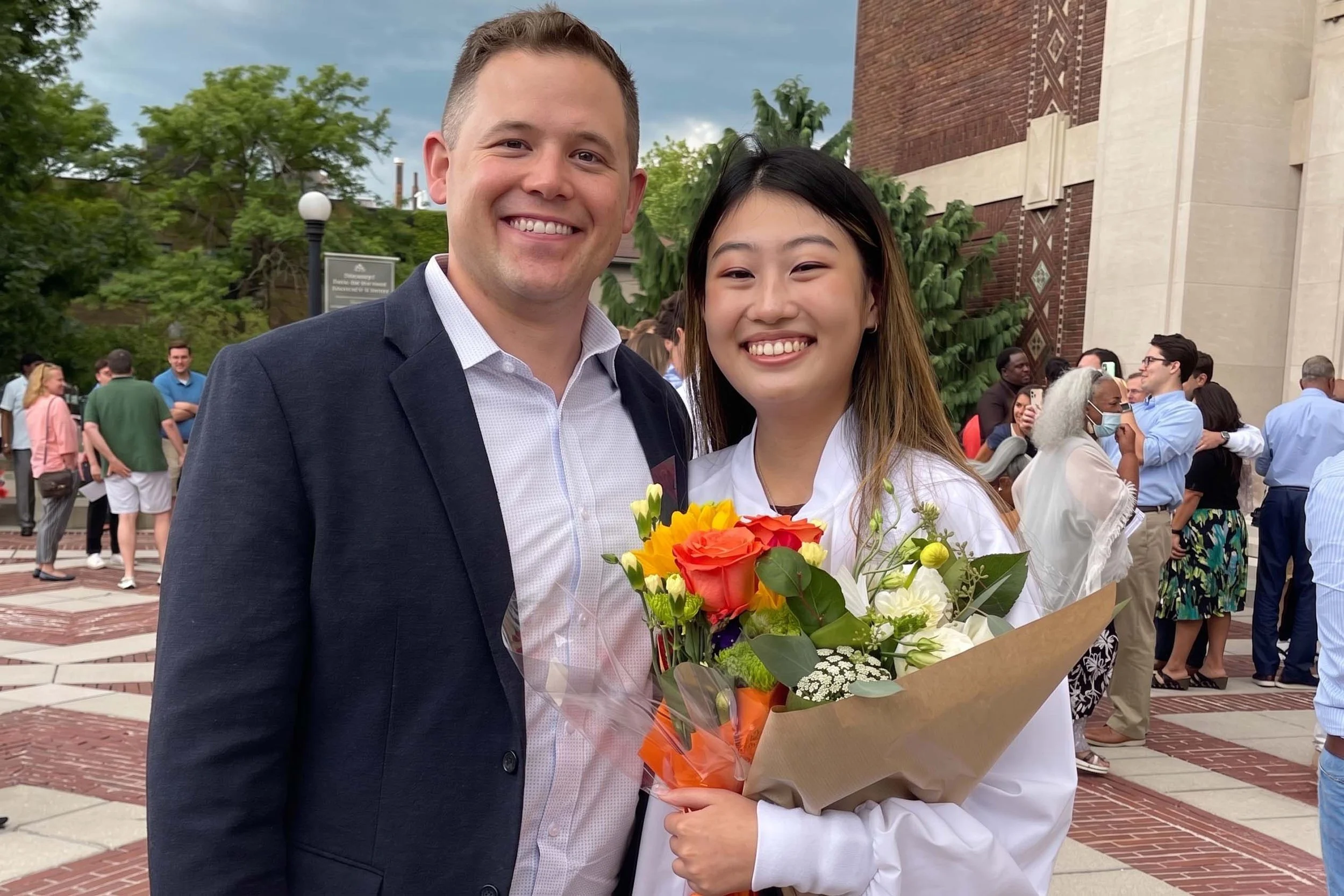 A young girl in a white outfit smiling and holding a bouquet of colorful flowers, standing next to a man in a dark suit outside on a cloudy day with other people gathered in the background.