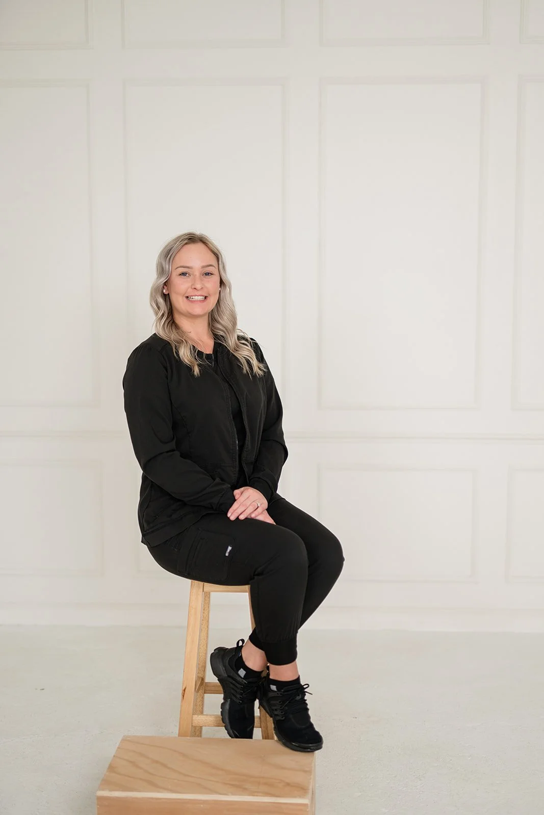 A smiling woman with blonde wavy hair sitting on a wooden stool against a plain off-white background.