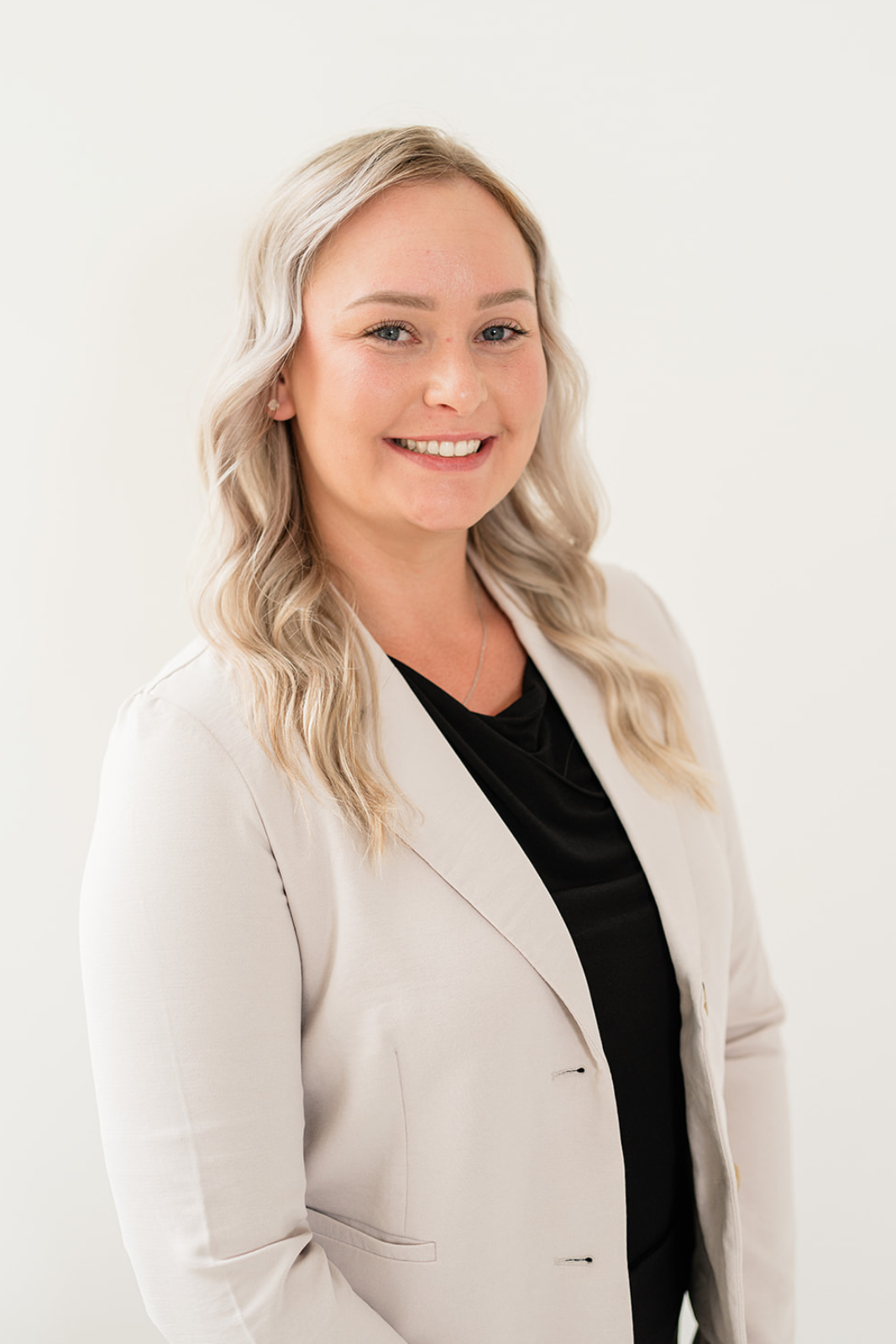 Professional woman with blonde hair styled in loose waves, smiling, wearing a beige blazer over a black top, standing against a white background.