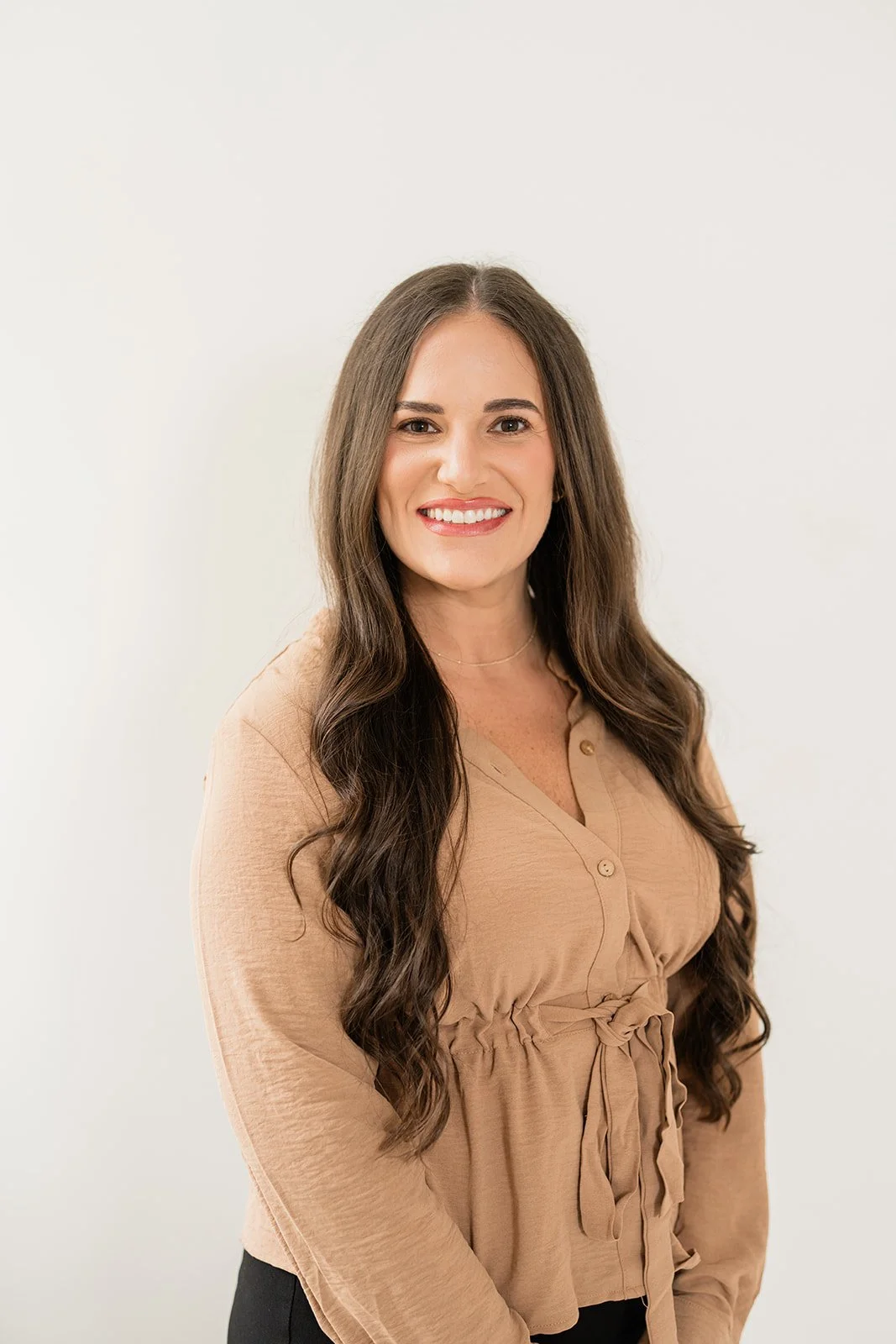 A woman with long wavy brown hair styled in loose curls, smiling, wearing a beige button-up blouse with a tied waist, standing against a plain white background.
