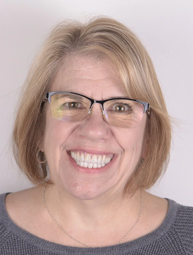 Close-up of a middle-aged woman with short, reddish-blonde hair smiling, wearing glasses and a gray top, against a plain white background.