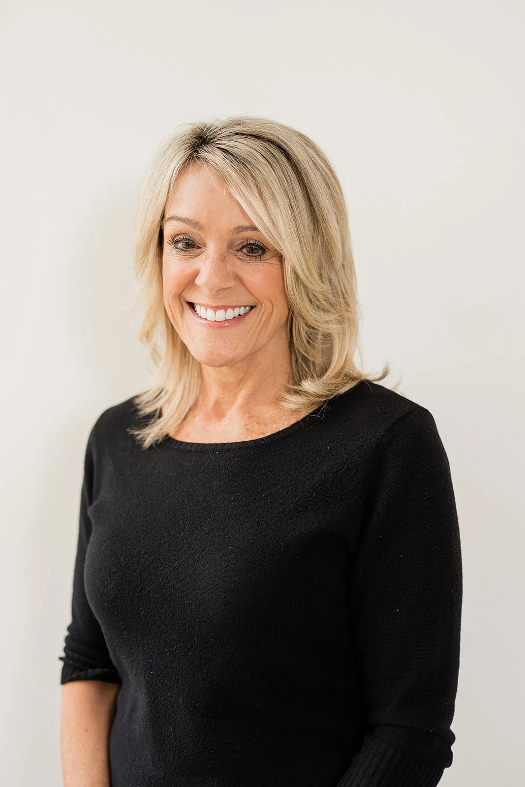 A woman with blonde hair smiling wearing a black top standing against a white background.