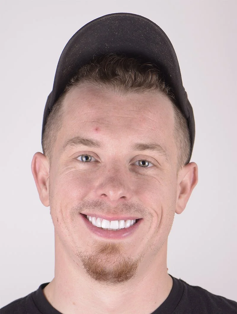 A young man with a light beard and mustache, wearing a black baseball cap backwards, smiling with straight white teeth, and dressed in a black shirt.
