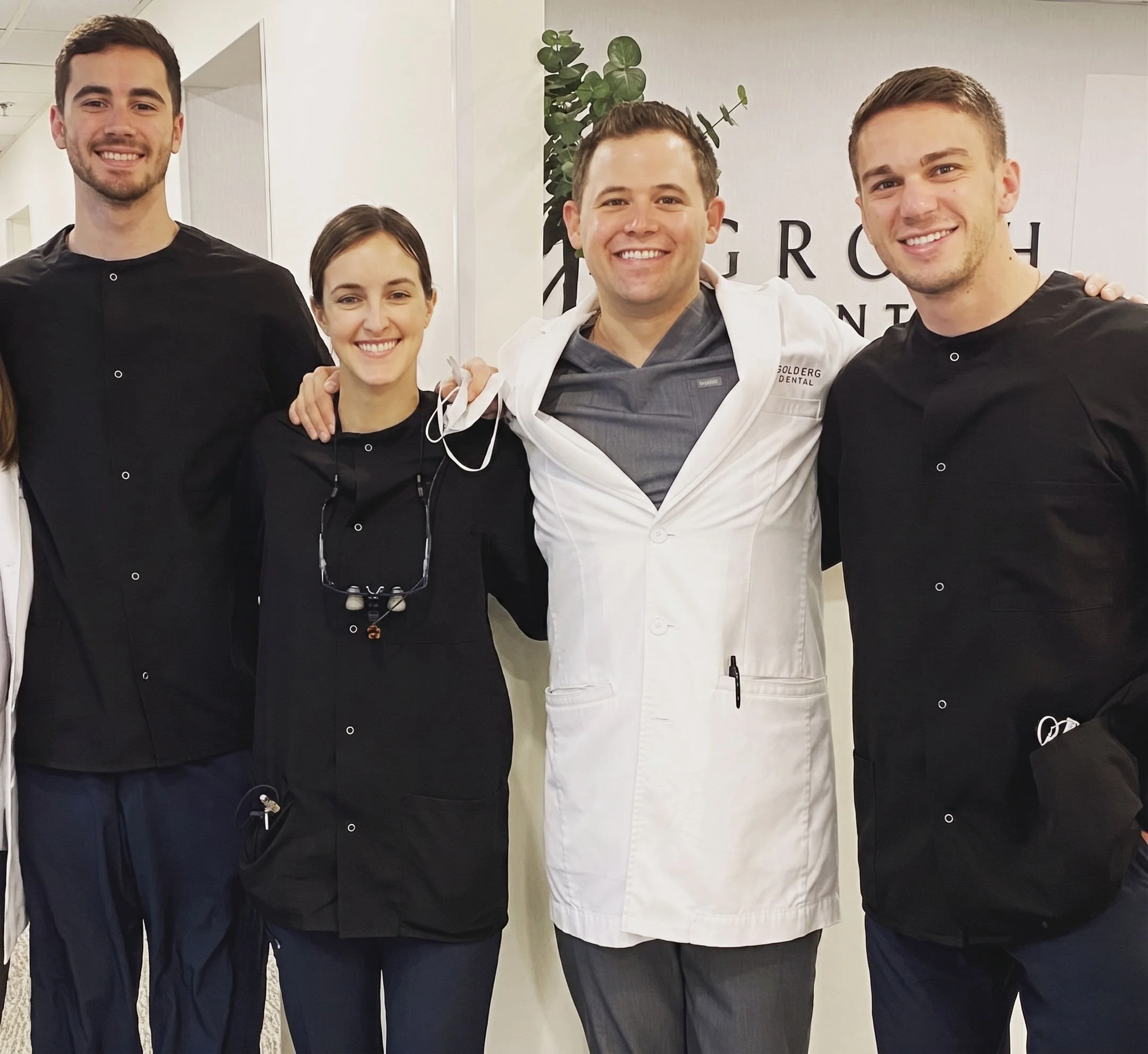 Four medical professionals, three men and one woman, standing together in a hospital or clinic setting nearest a wall with the words 'GOLDERG DENTAL' and a leafy plant, smiling at the camera.
