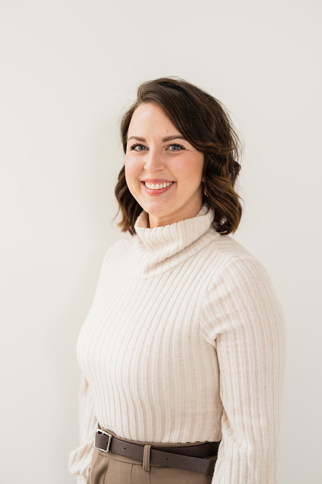 A woman with shoulder-length wavy dark hair, smiling, wearing a cream-colored turtleneck sweater and beige pants, standing against a plain white background.