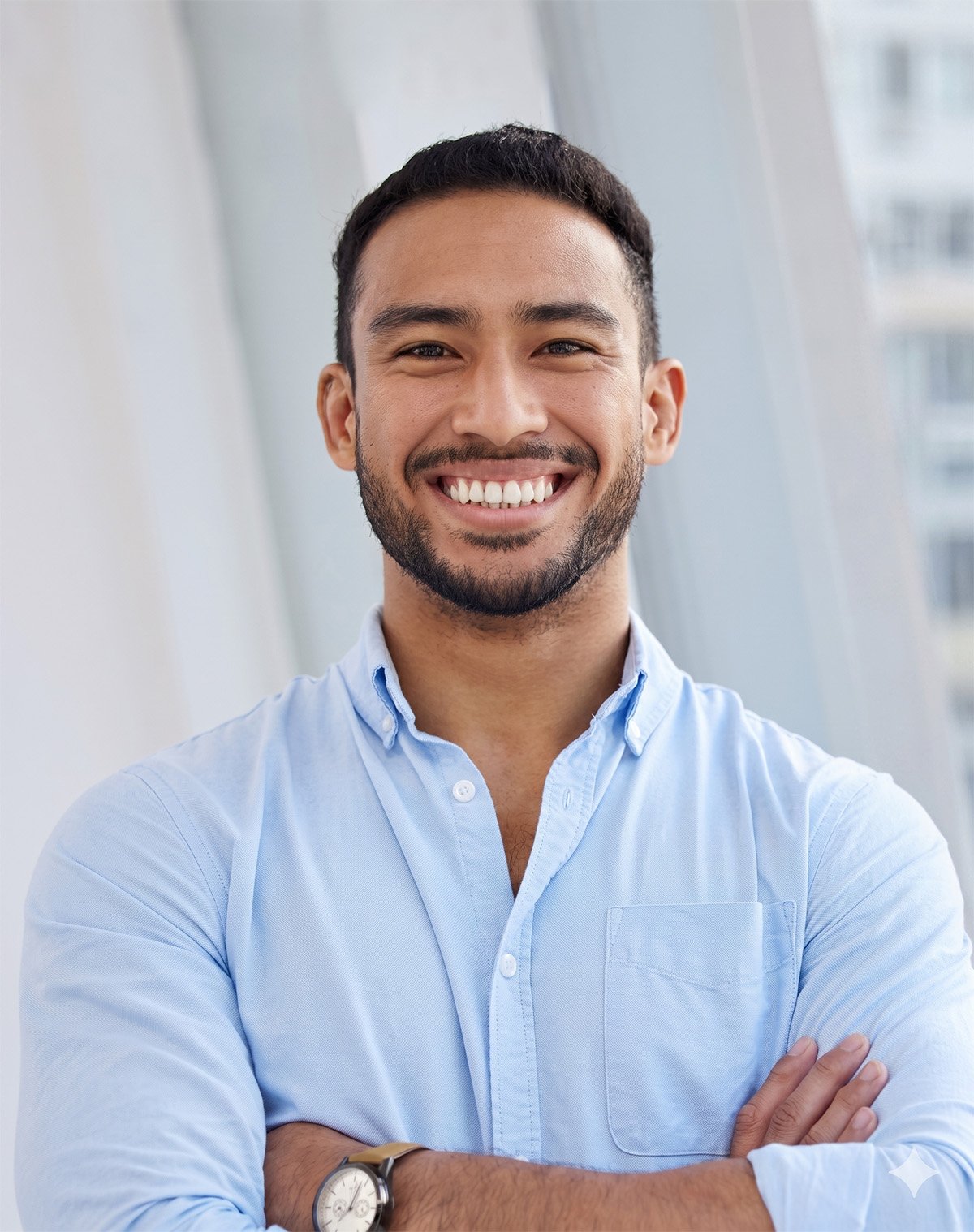 A smiling man with dark hair and a beard, wearing a light blue button-up shirt, standing with arms crossed.