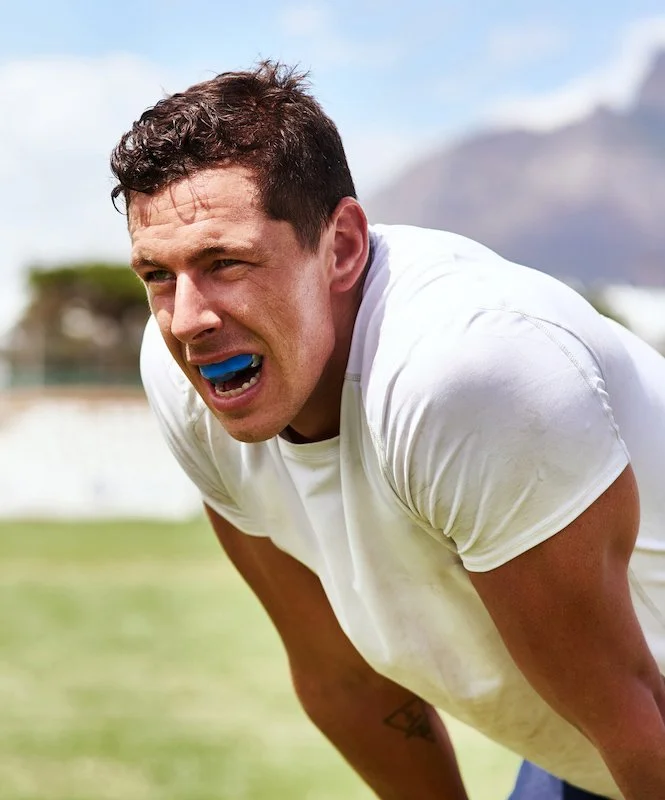 Male athlete in a white shirt with a blue mouthguard, leaning forward with an expression of exertion or pain, on a grassy field with mountains in the background.
