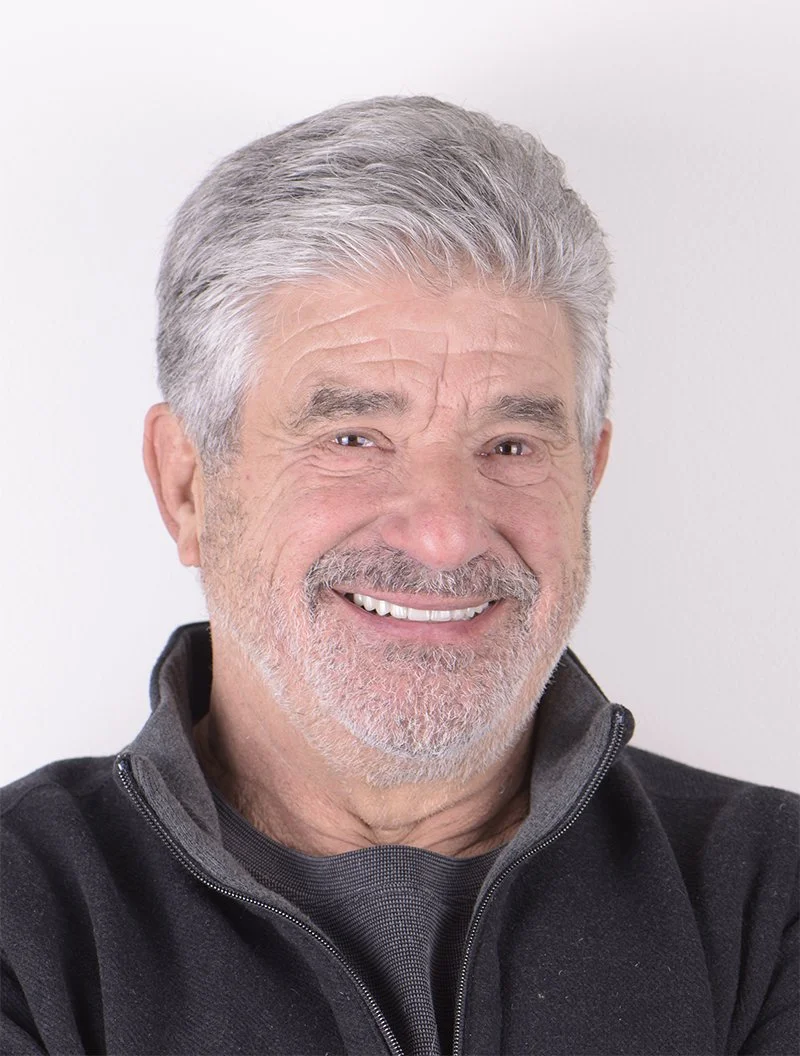 Close-up of an older man with gray hair and a beard, smiling, wearing a black zip-up jacket against a white background.