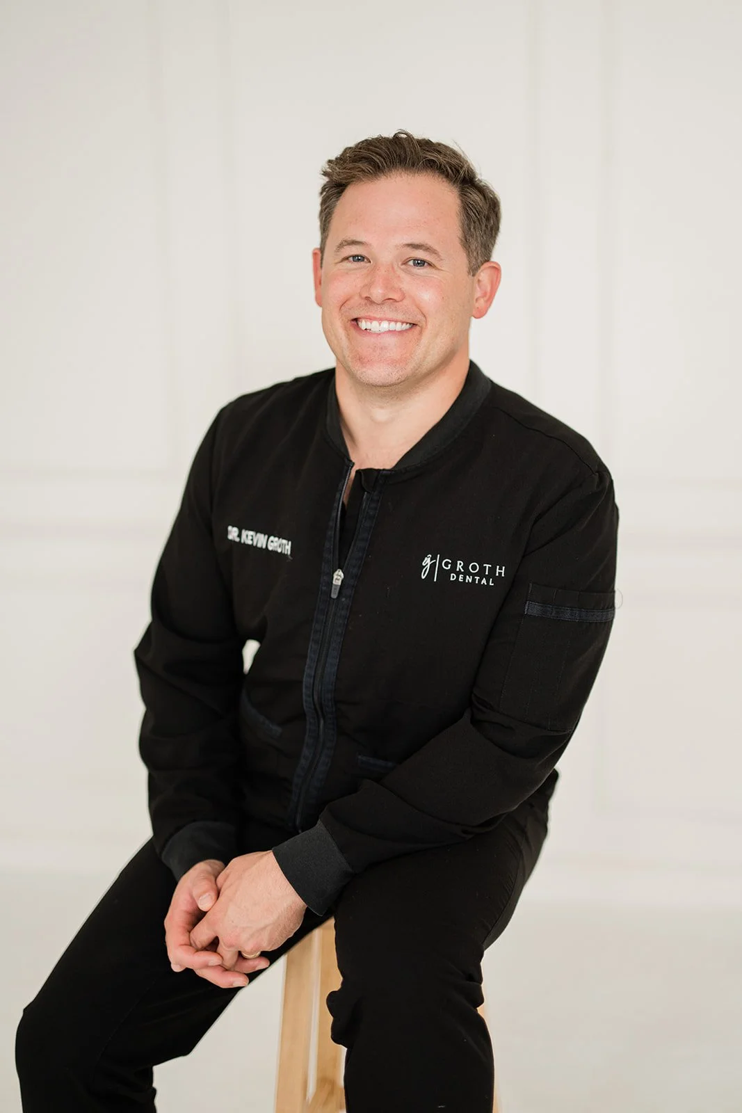 Portrait of a smiling man in a black dental professional uniform, sitting on a stool in front of a plain light-colored background.