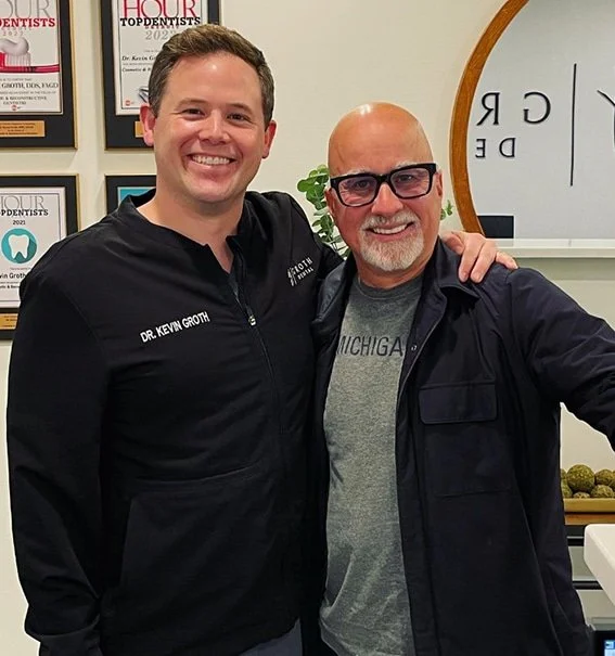 Two men smiling and standing close together in an office or clinic. The man on the left is wearing a black jacket with Dr. Kevin Groth's name embroidered, and the man on the right is wearing glasses and a gray T-shirt with a black jacket. There are framed awards or certificates and a whiteboard in the background.