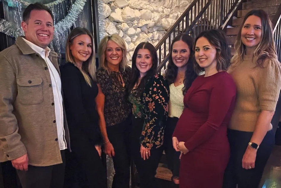Seven smiling people posing together at a social gathering indoors, with a stone wall and staircase in the background.