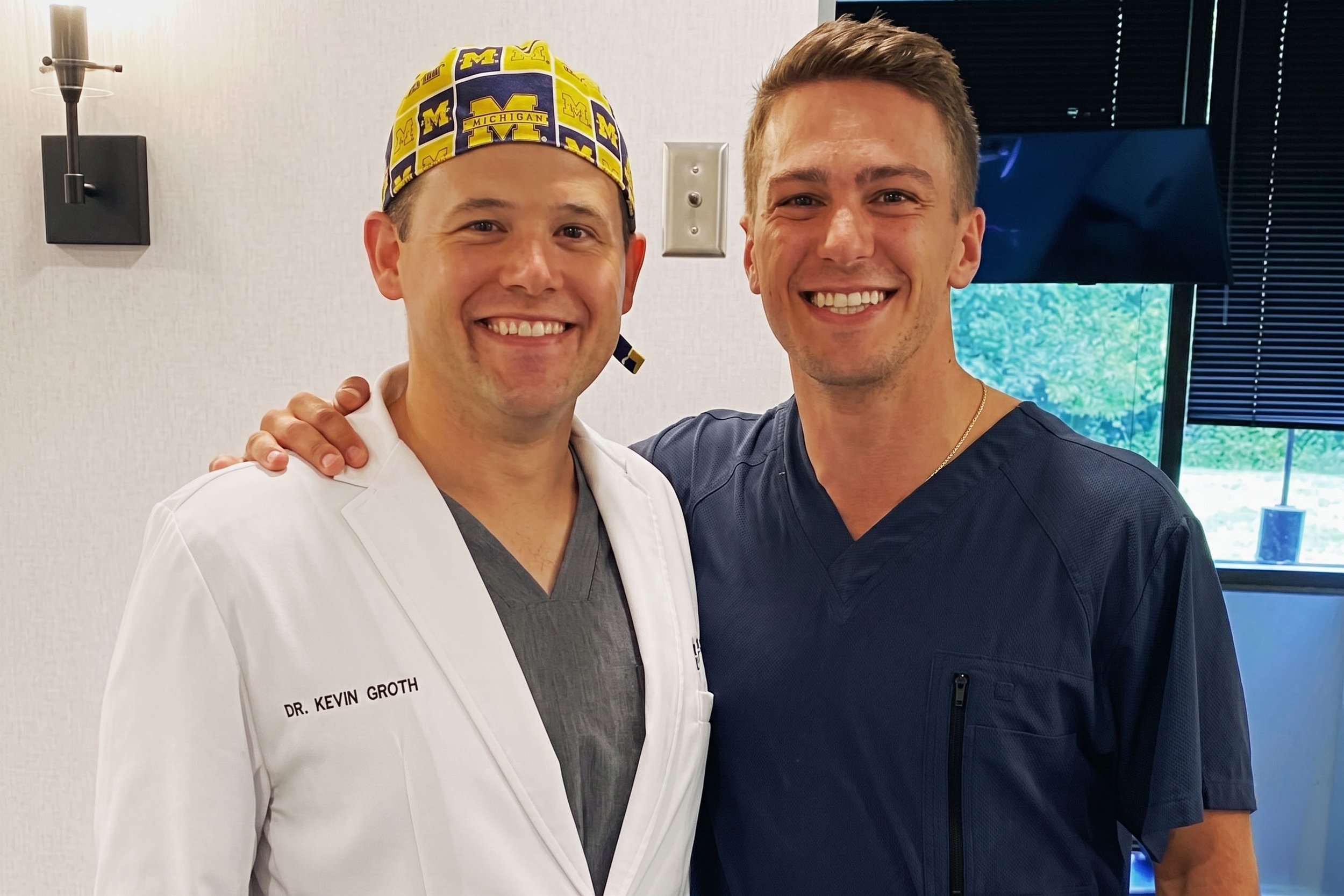 Two smiling men standing close together, one in a white lab coat with a name tag that reads 'Dr. Kevin Groth' and a Michigan-themed scrub cap, and the other in a dark medical scrub top. They are in a room with a window and blinds in the background.