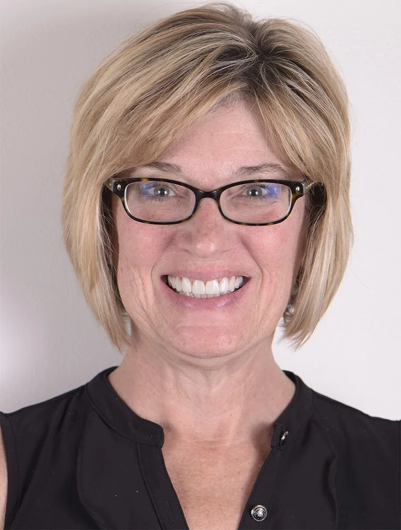 A smiling woman with blonde hair and black glasses, wearing a black sleeveless top, standing against a plain light background.
