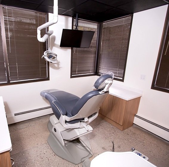 Empty dental examination room with dental chair, overhead light, monitor, and window blinds.