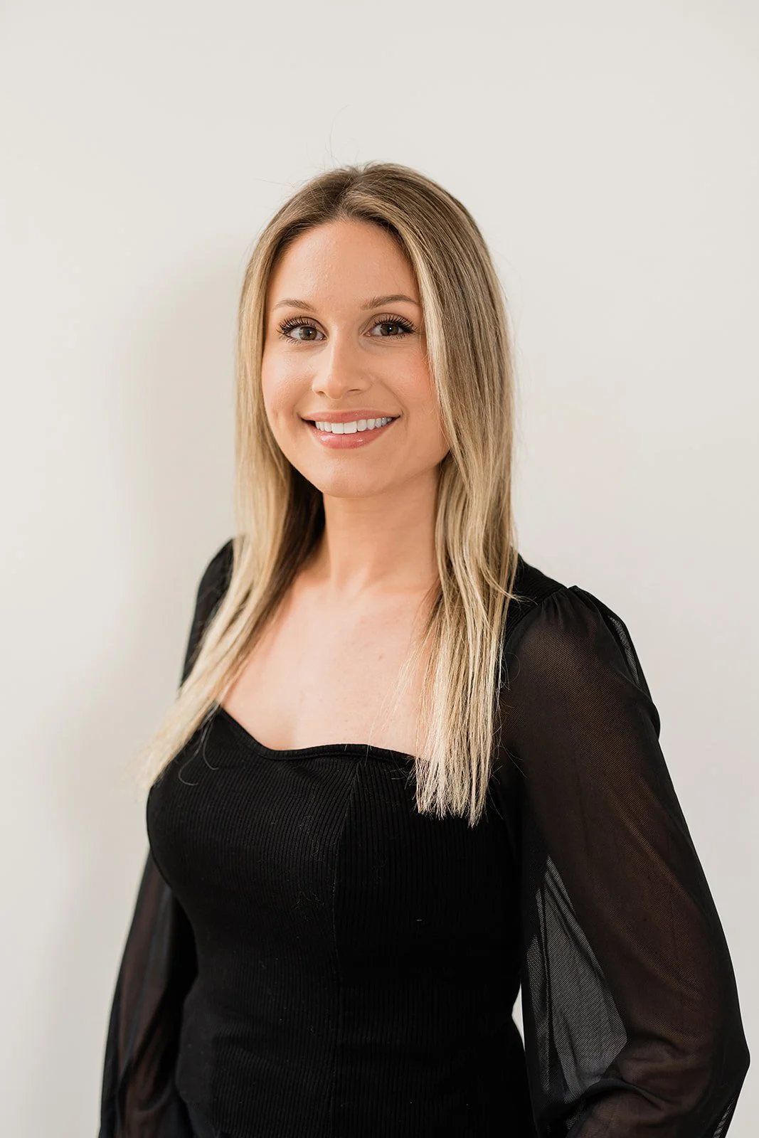 A woman with long blonde hair smiling at the camera, wearing a black top with sheer sleeves, standing against a plain white background.