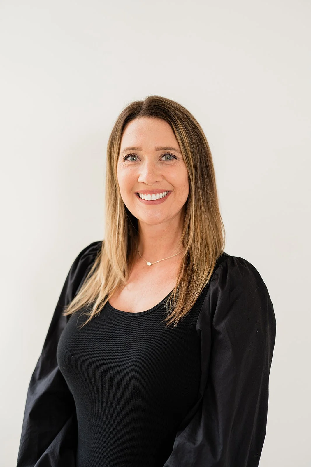 A smiling woman with long, light brown hair, wearing a black top and a delicate necklace, standing against a plain white background.