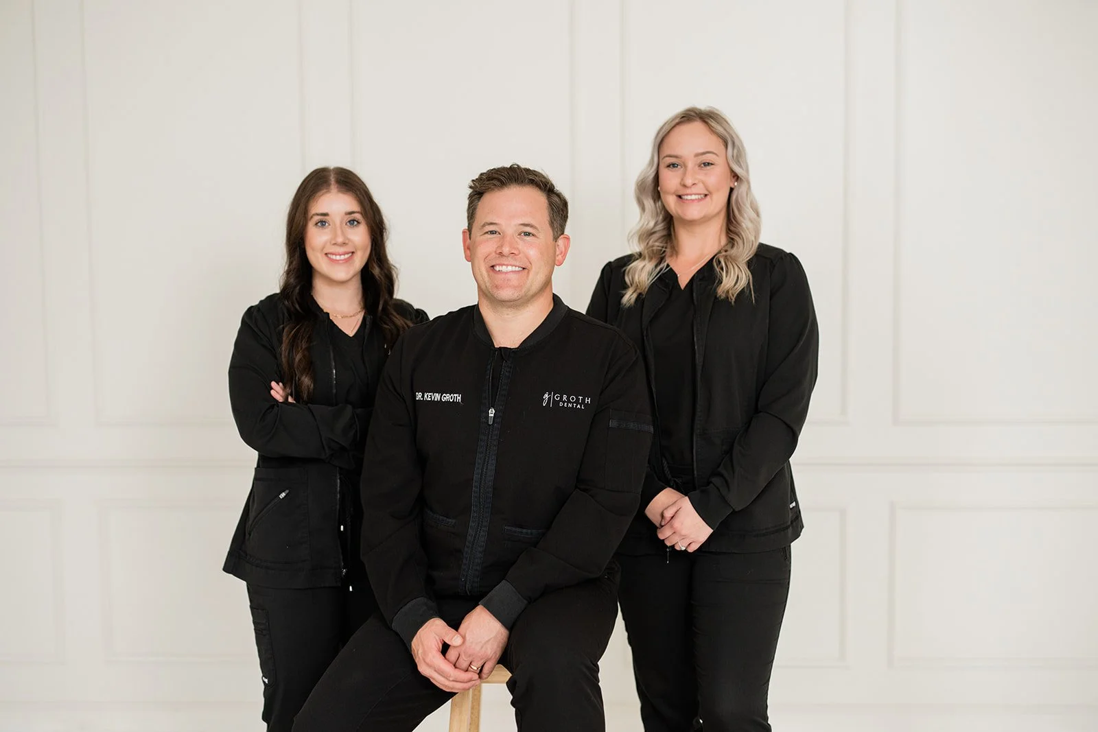 Three women and one man posing for a photo, dressed in black, in front of a white wall. The man in the front is wearing a black jacket with 'Dr. Kevin Groth' and 'Groth Dental' on it, seated on a stool. The women are standing behind him, smiling.