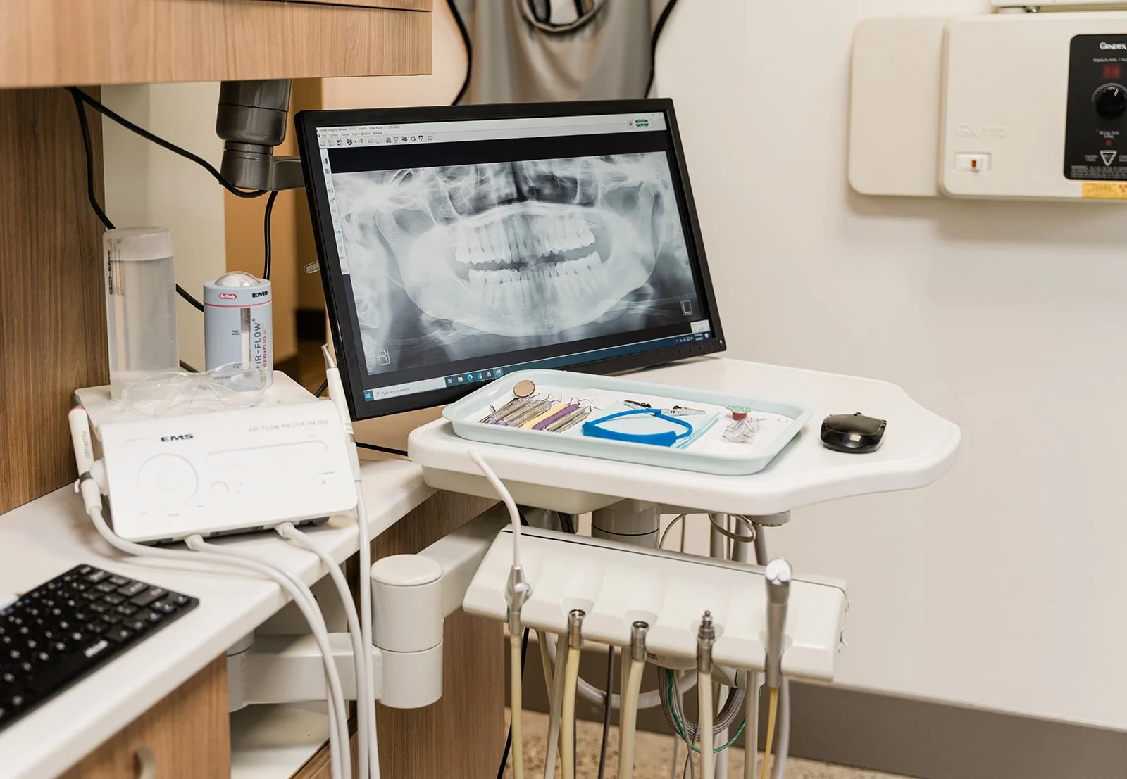A dental office with a monitor displaying a panoramic dental X-ray, dental tools on a tray, and various dental equipment and supplies on counters.