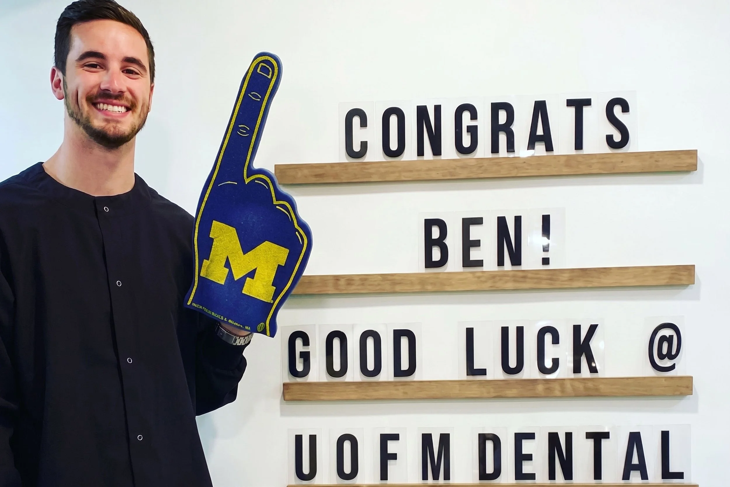 A young man smiling while holding a foam finger with the University of Michigan logo, standing in front of a wall display with congratulatory messages, including one that says 'CONGRATS' and another with added text that says 'HOPE, GOOD LUCK, & MATER