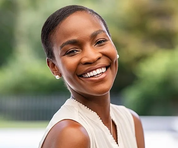 A smiling young woman with dark skin, wearing pearl earrings and a sleeveless white top, outdoors with a blurred green background.