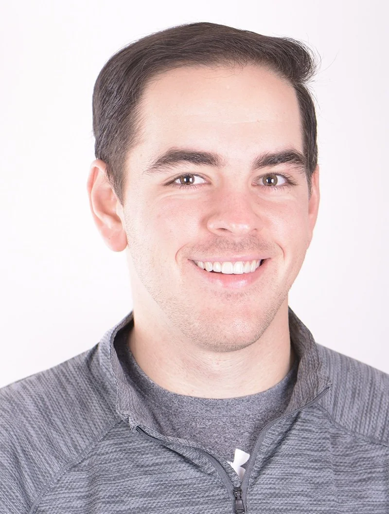 Portrait of a young man with brown hair and gray eyes, smiling, wearing a gray zip-up athletic jacket against a white background.