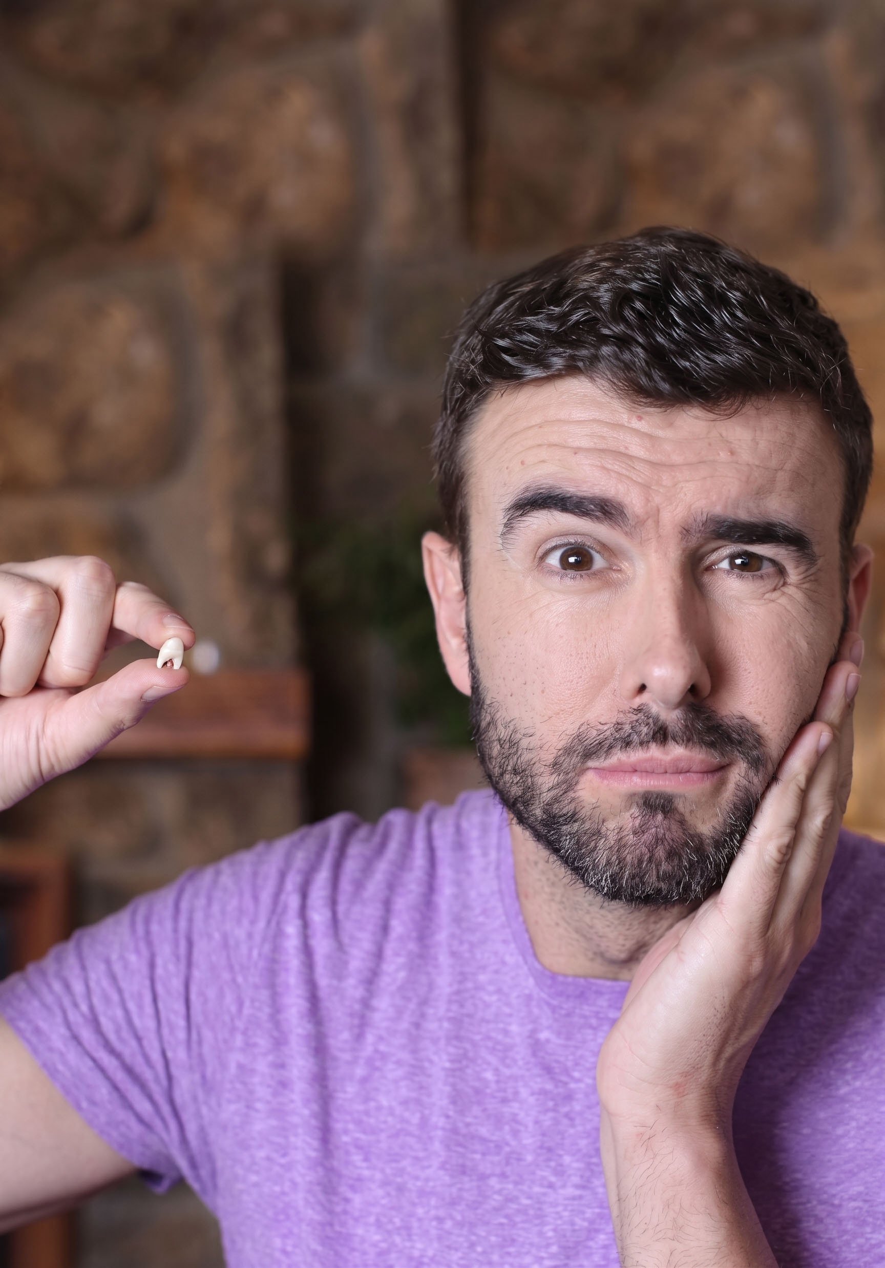 Close-up of a man with curly dark hair and a beard wearing a purple shirt, holding a piece of jewelry, with a blurred brick wall background.