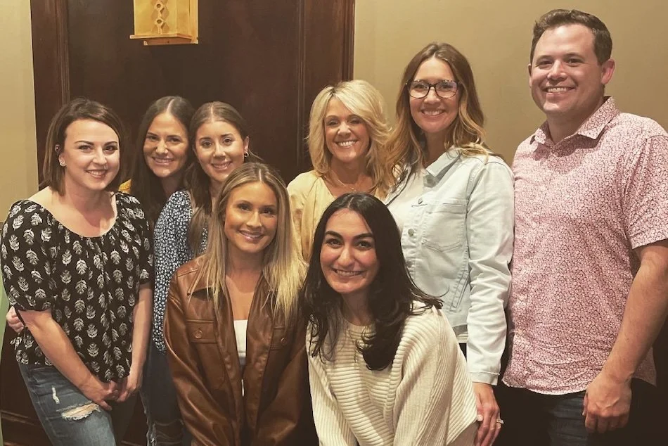 Group of nine smiling people, including women and one man, gathered indoors for a photo.