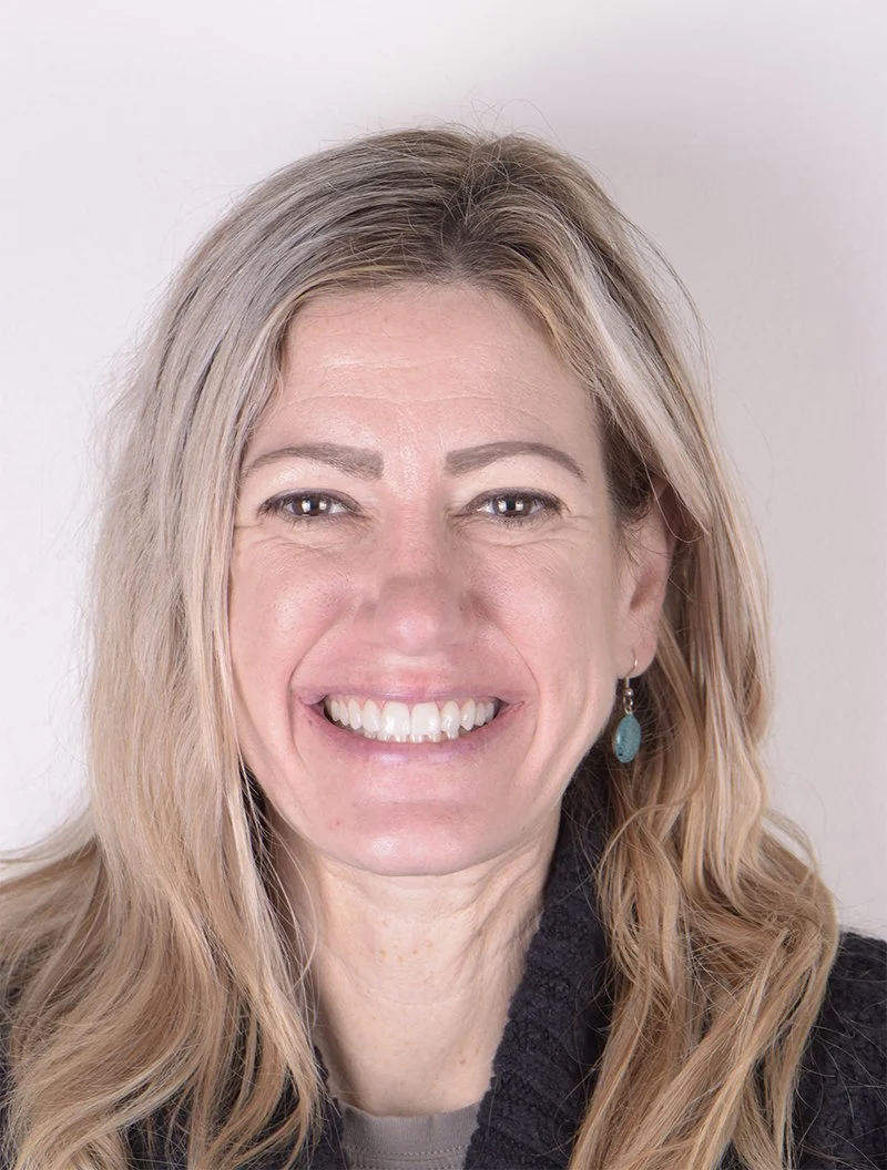 A smiling woman with long, wavy blonde hair, wearing a black sweater and turquoise earrings.