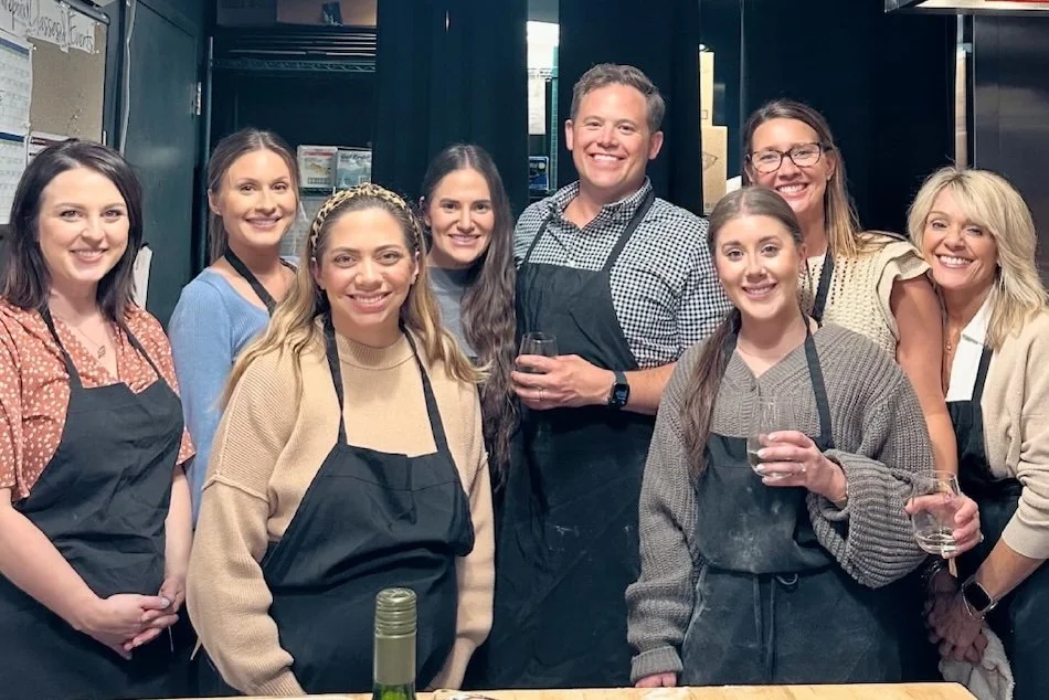 Group of eight people, six women and two men, smiling and holding drinks, wearing aprons, standing in a kitchen or food service setting.