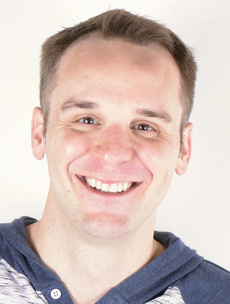 Close-up of a smiling young man with short brown hair, wearing a blue hoodie with a gray shirt underneath, against a plain white background.
