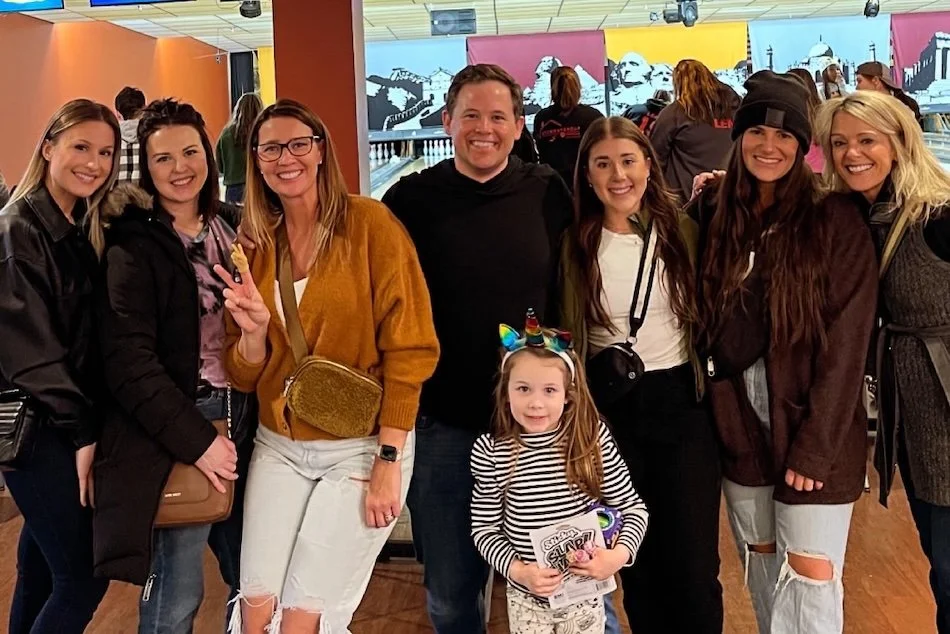 A group of eight smiling people, including a young girl with a unicorn headband, posing together indoors with colorful artwork and people in the background.