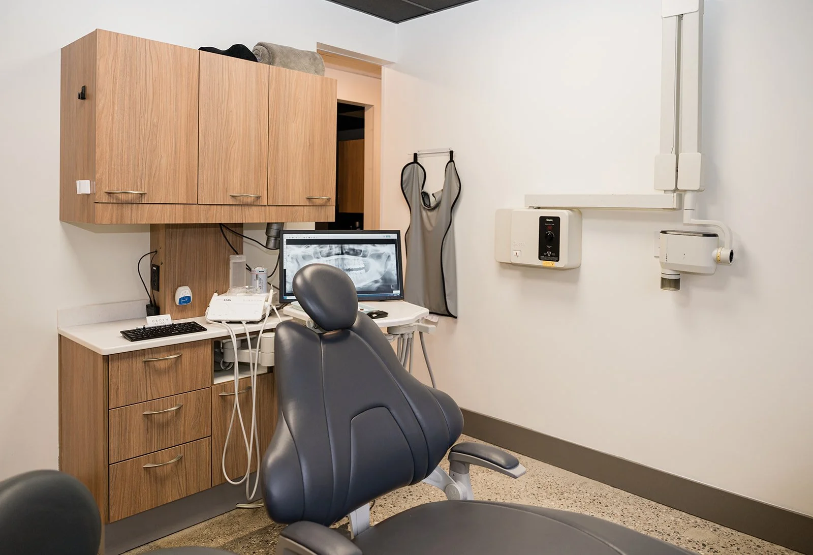 Dental examination room with dental chair, computer monitor displaying an X-ray, wooden cabinetry, and dental equipment.