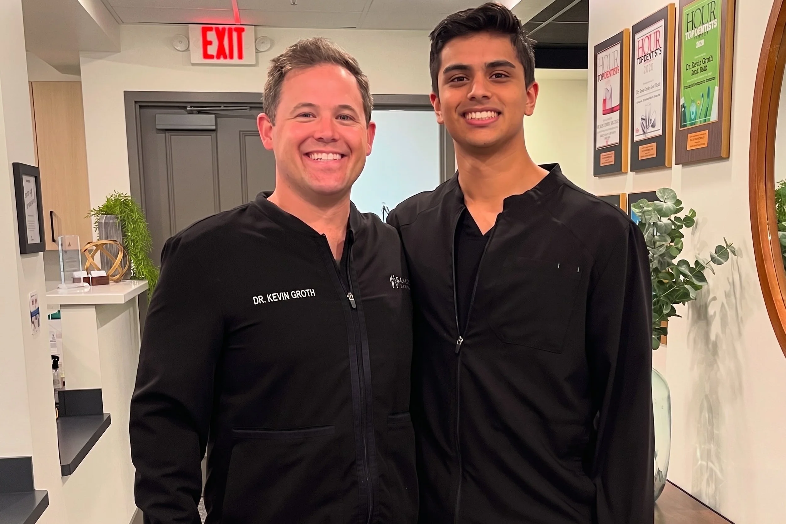 Two smiling men in black medical scrubs standing side by side in a modern office or clinic, with framed awards and plants in the background.