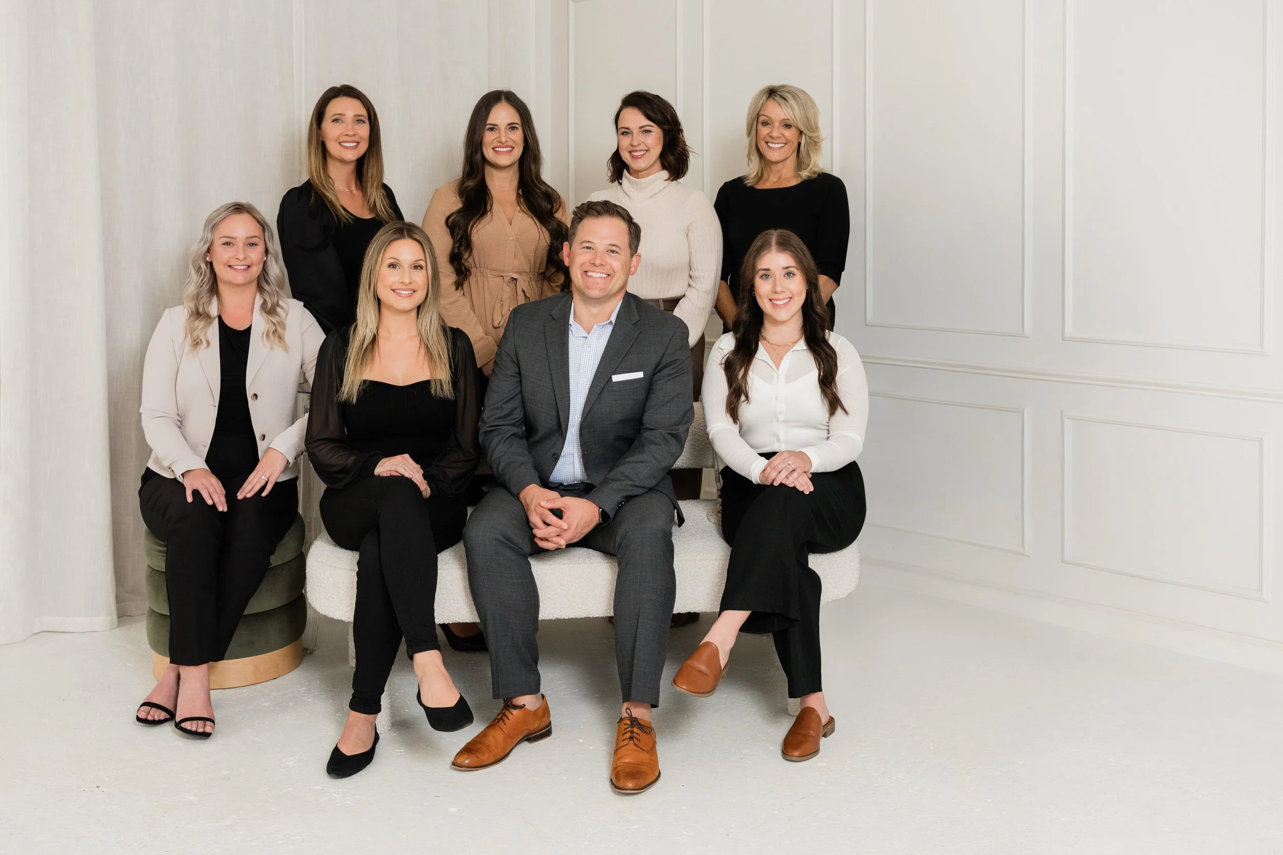 Group of ten adults, eight women and two men, posing together in a professional setting, some sitting and some standing, smiling at the camera.