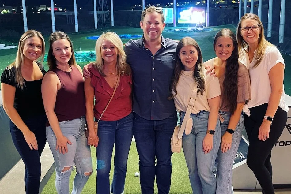 Seven people, five women and two men, standing together on an indoor mini-golf course, smiling at the camera.