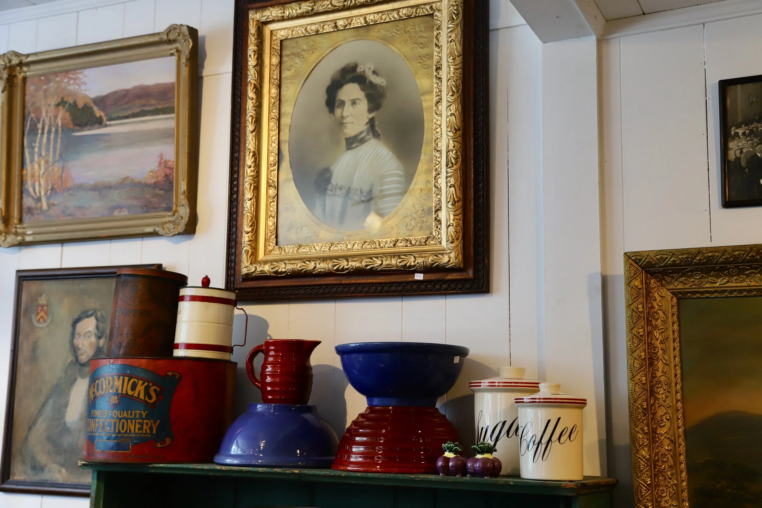 A collection of vintage and decorative items on a green shelf and wall, including framed paintings, a black and white portrait of a woman, ceramic vases, bowls, and tins.