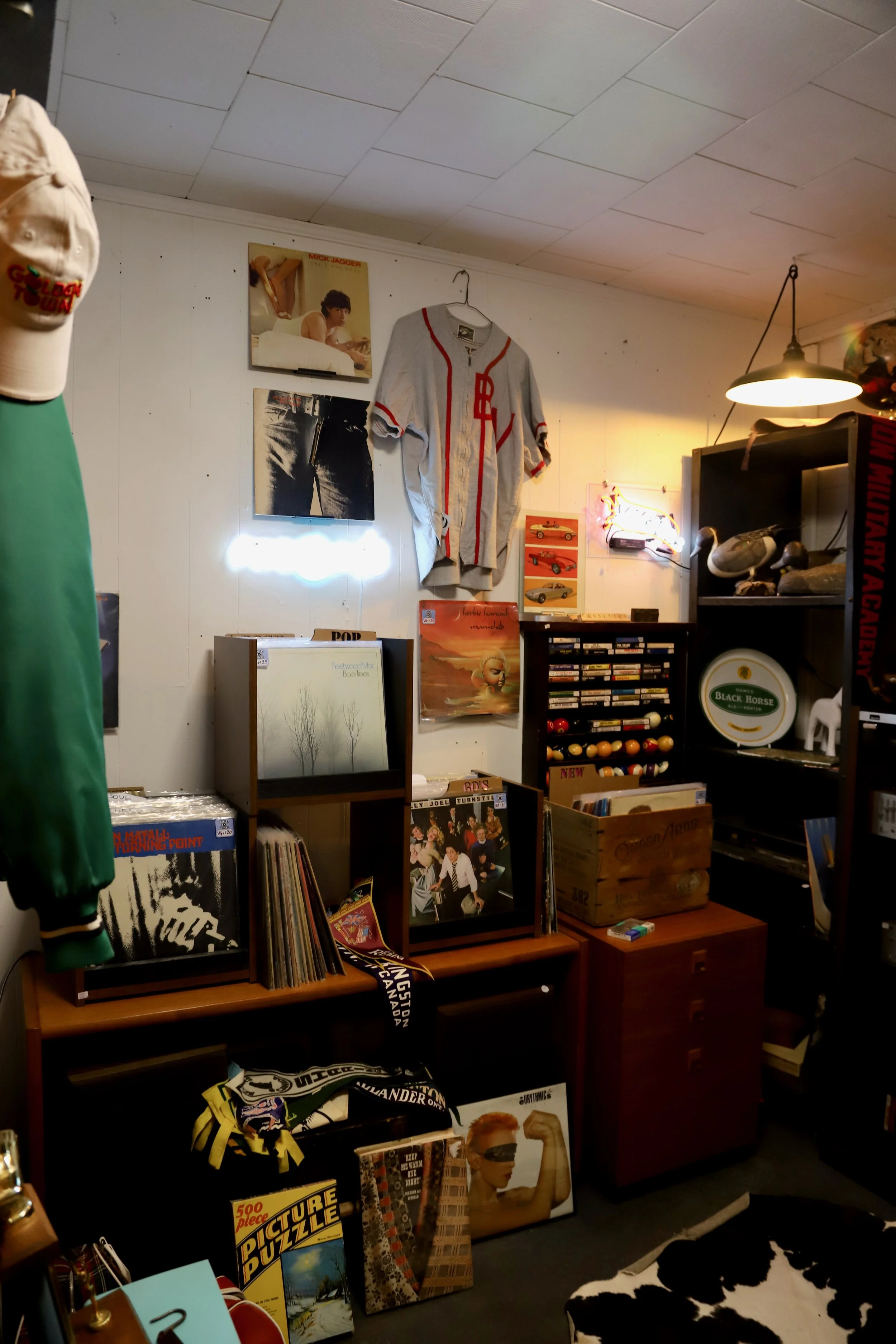 A room filled with vintage albums, posters, and collectibles, including a baseball jersey hanging on the wall, a black shelf with small toys and items, and books and puzzles stacked on surfaces. The ceiling is white with tiles, and there's a cowhide rug on the floor.