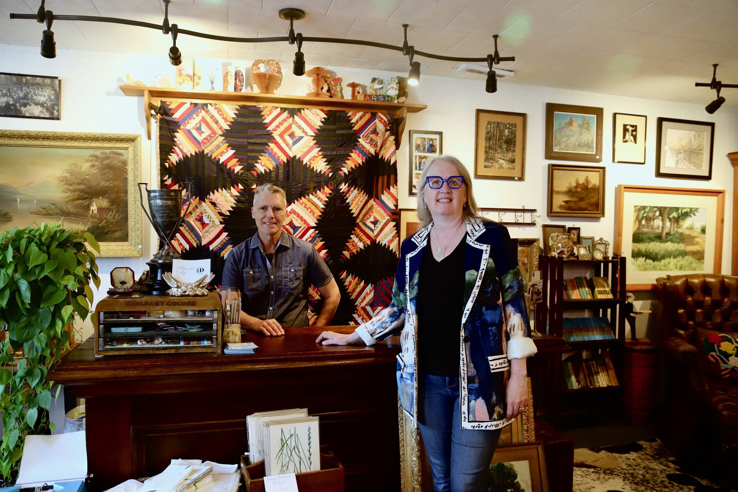 A woman with blonde hair, glasses, and a colorful jacket stands in front of a man seated behind a desk, both smiling at an art gallery or antique store. The background features framed paintings and artwork, and a quilted wall hanging. The woman is resting her hand on the desk.