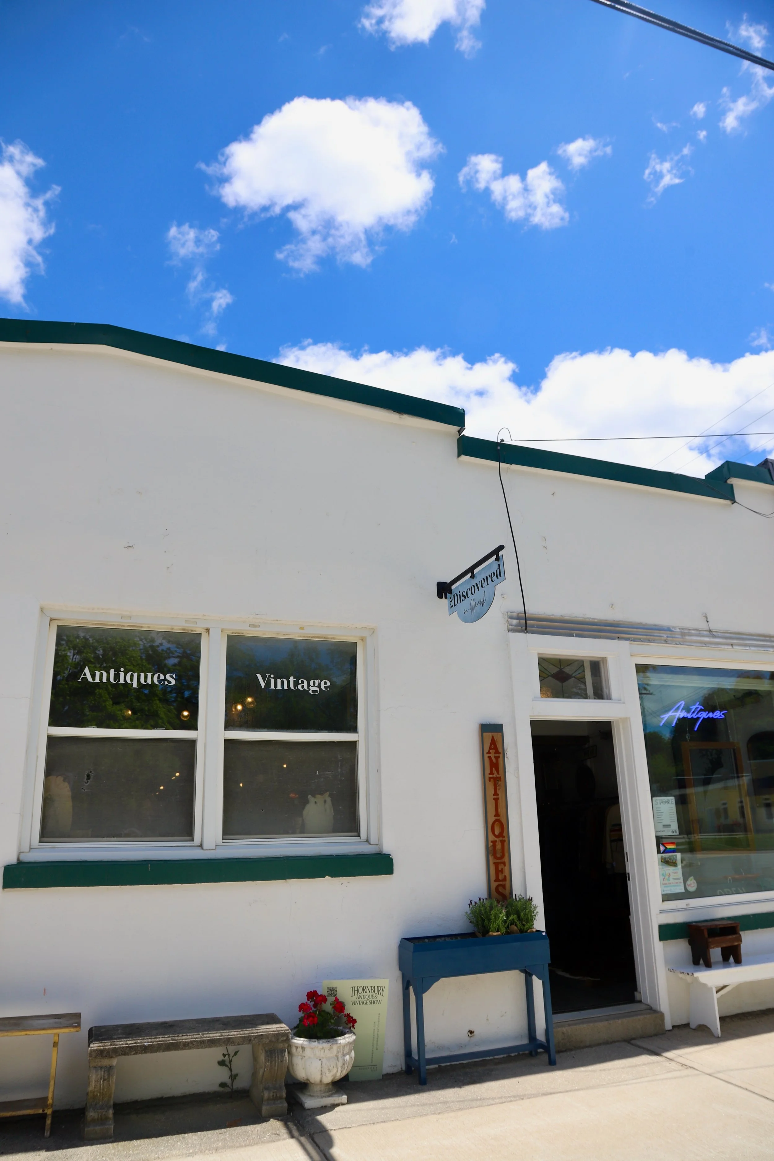White building with large window displaying the words 'Antiques' and 'Vintage' on the glass. Exterior decor includes a wooden bench, a white flower pot with red flowers, a blue table with a green plant, and a vertical sign that reads 'Antiques' in red letters. The sky is blue with a few white clouds.