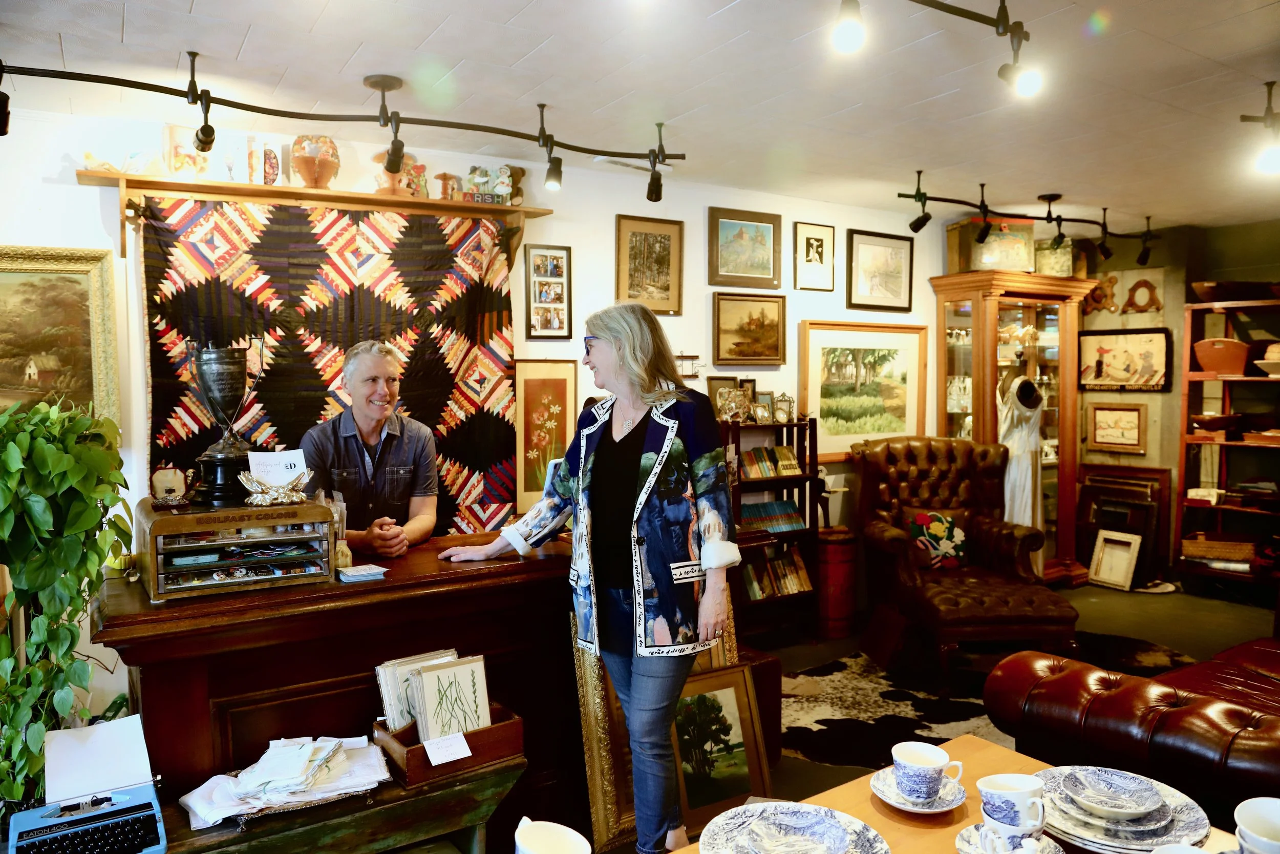 Interior of an art gallery or antique shop with framed paintings and decorative items on the walls and shelves. Two women appear to be engaged in conversation near a wooden counter, surrounded by various artworks, ceramics, and collectibles.