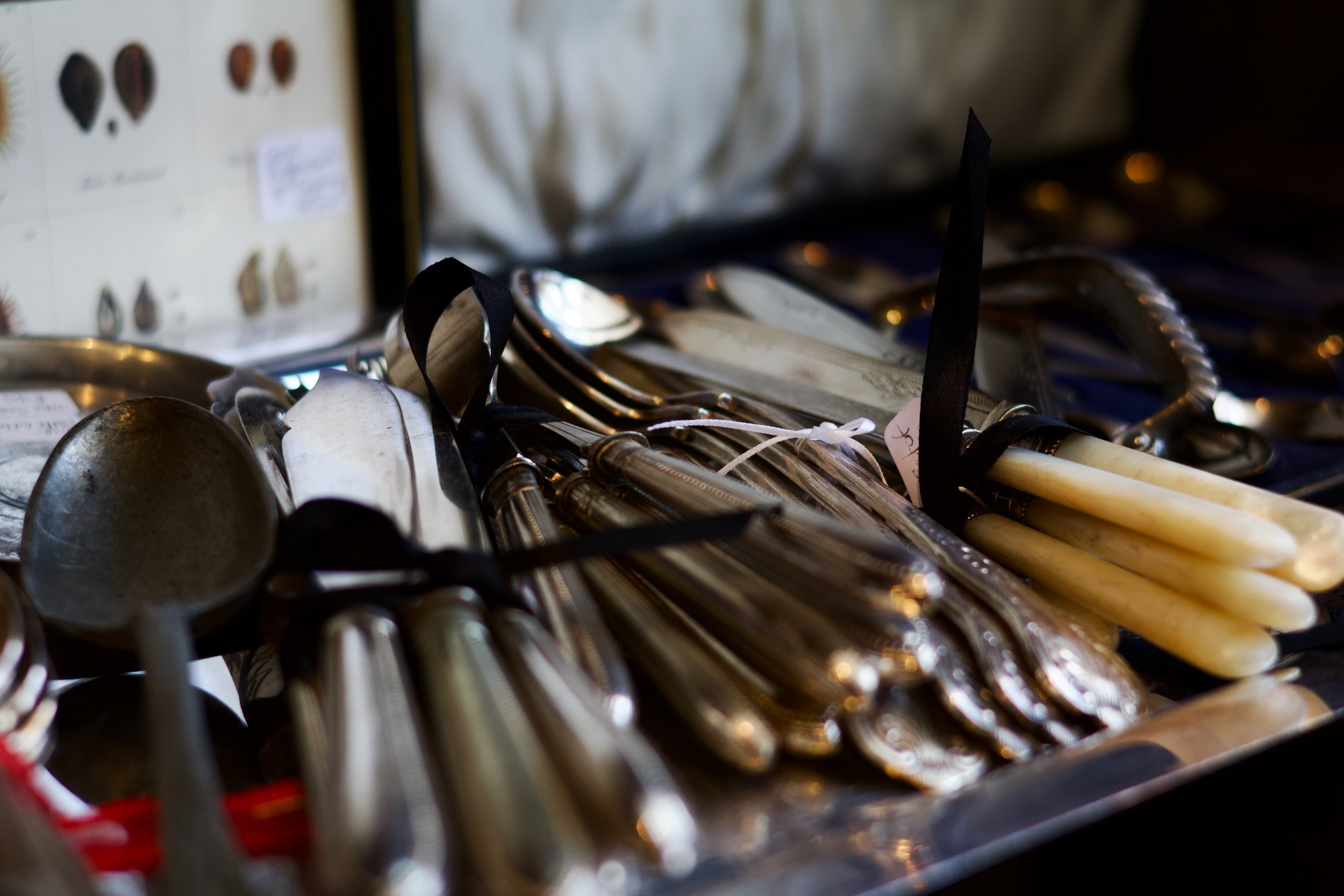 A collection of vintage silverware including spoons and forks arranged on a surface, with a blurred background showing a display of various earrings.