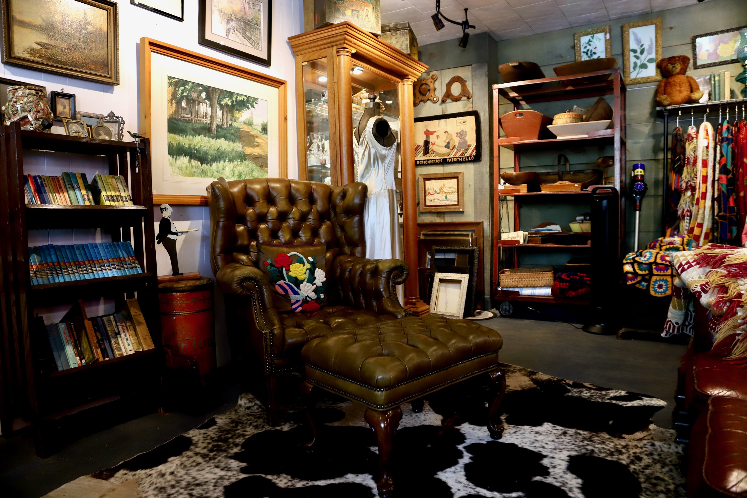 A cozy vintage-style room with walls decorated with framed artwork and shelves filled with books and decorative items. A leather tufted armchair sits on a black and white cowhide rug, with colorful crochet pillows. There are many various shelves and picture frames around the room, creating a warm and eclectic atmosphere.