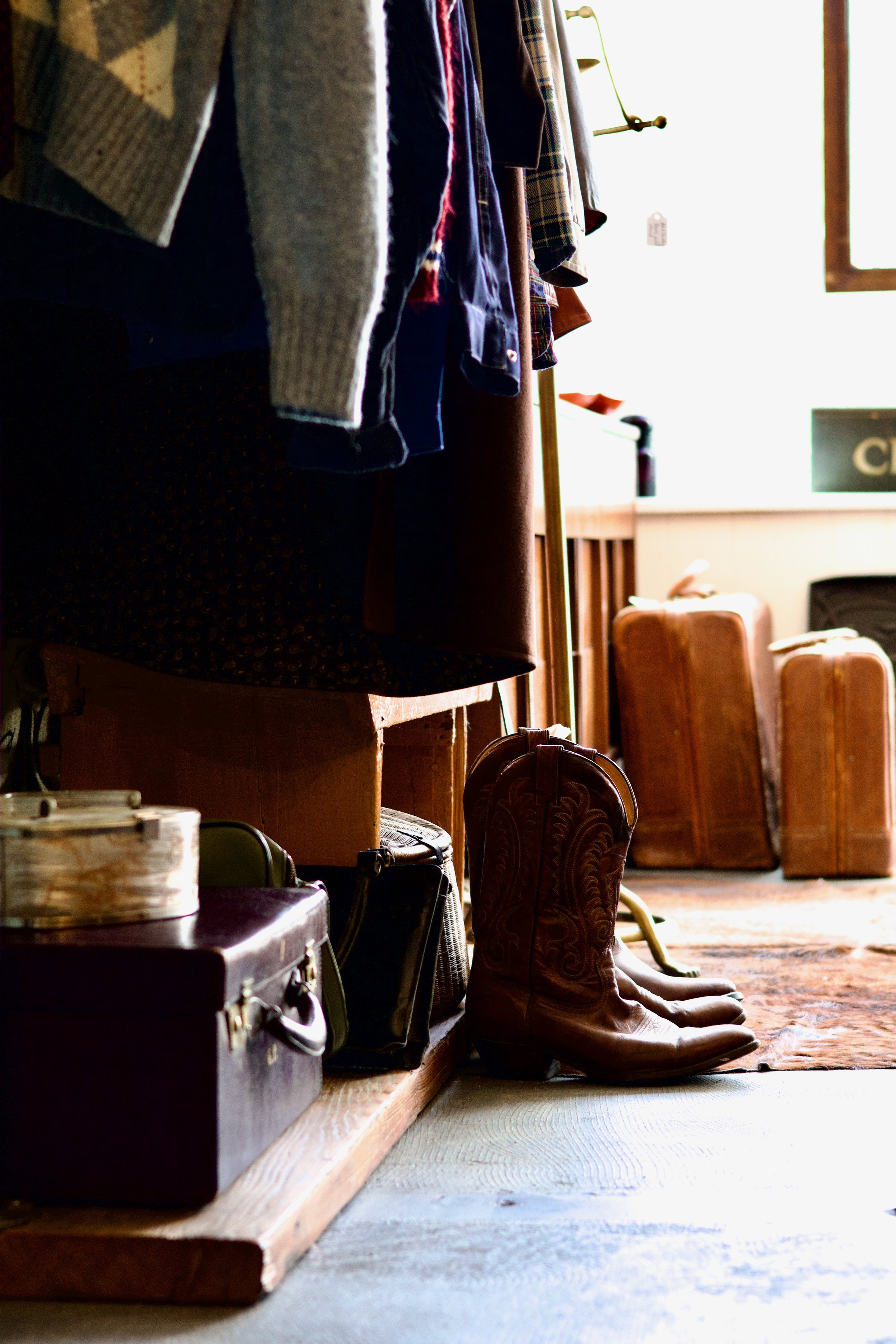 Vintage cowboy boots and suitcases placed on a wooden ledge, with hanging clothes above and leather chairs behind, in a cozy vintage shop or dressing room.
